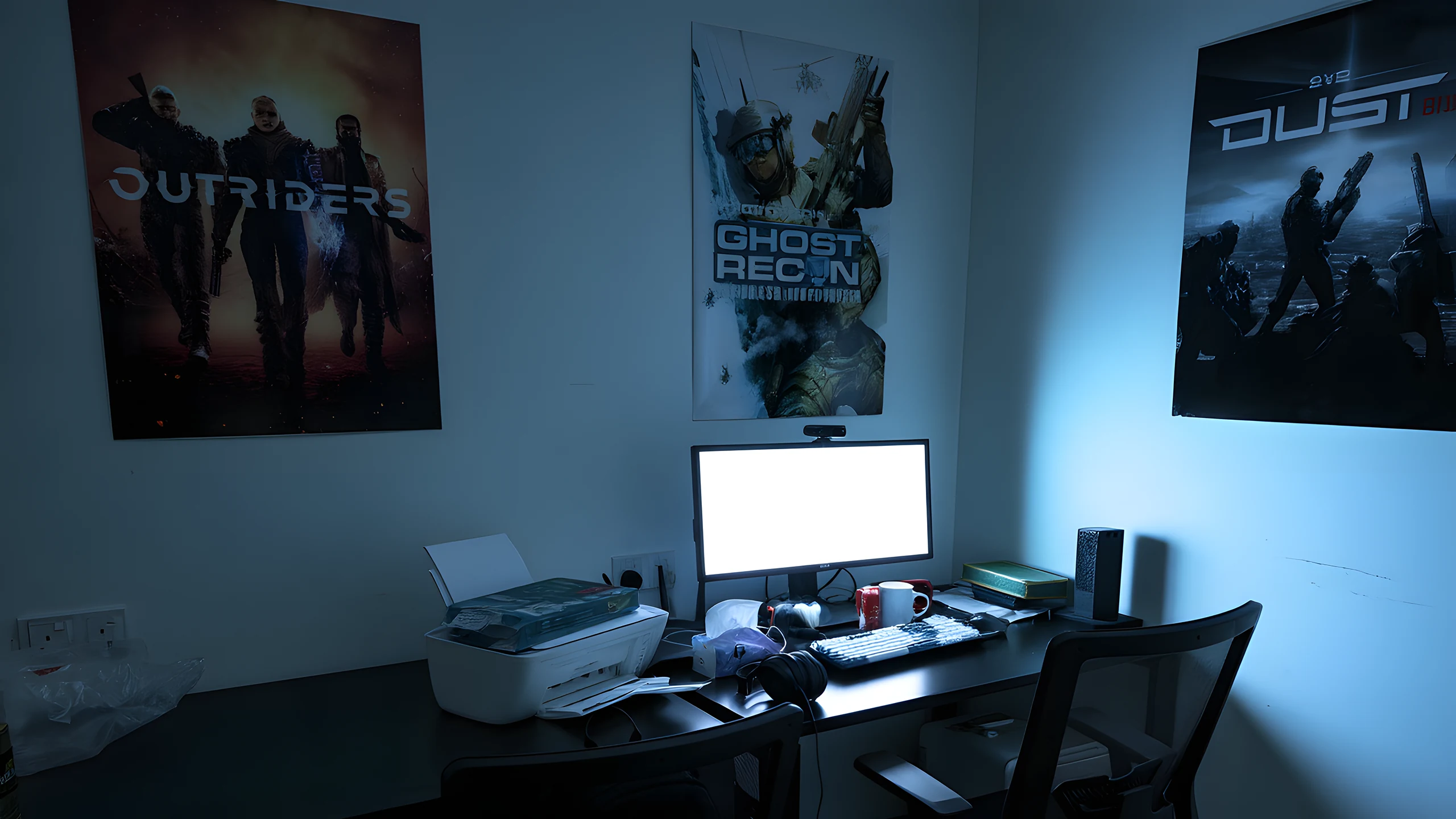 A desk in low blue light with a monitor, keyboard, headphones and a printer on it, and framed posters on the wall behind.