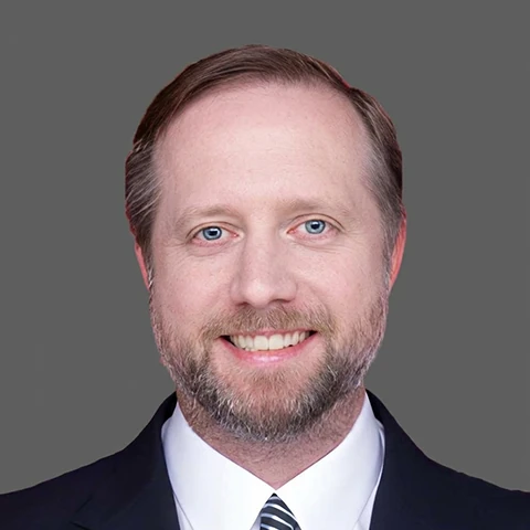 A man in a dark suit jacket and a diagonally striped tie, smiling at the camera against a plain grey background.