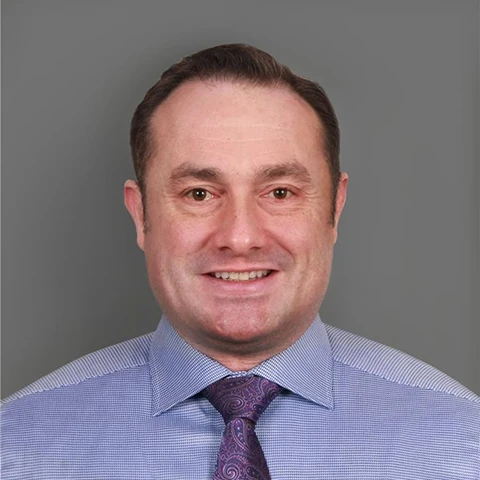 A man in a light blue patterned shirt and a purple patterned tie, smiling at the camera against a plain grey background.